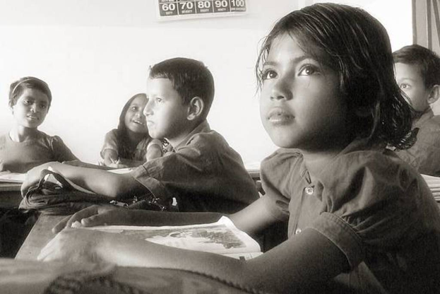 Eine Gruppe von Menschen an Tischen mit Büchern und Taschen, ein Kalender mit Zahlen und Text an der Wand und eine Tür oben rechts.