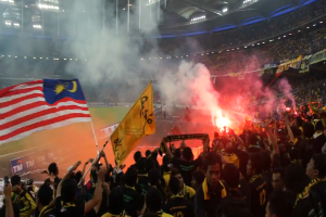 Eine Menschenmenge in einem Stadion mit Fackeln, Fahnen und einer Kamera, mit Rauch und Schildern, die "Malaysia vs. Malaysia Live-Streaming" lesen.