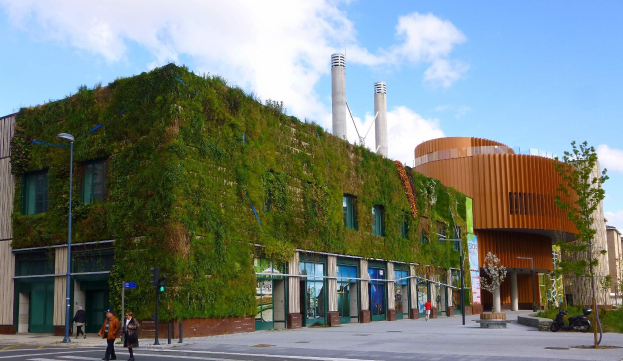 Große nachhaltige Gebäude mit einer Green Wall, umgeben von Straßeninfrastruktur, Fahrzeugen, Füpfölern, Bäumen und einem bewölktem Himmel.