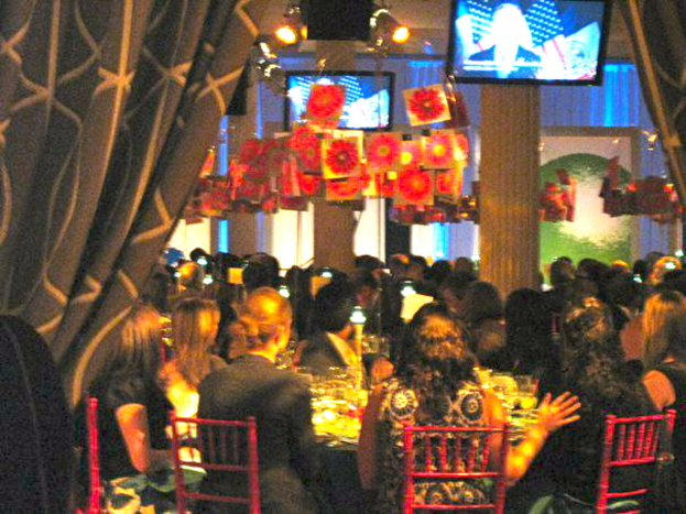 Eine Gruppe von Menschen sitzt an Tischen mit Essen, Fernsehgeräten, Lampen, hängenden Papieren und Vorhängen in einem Raum.