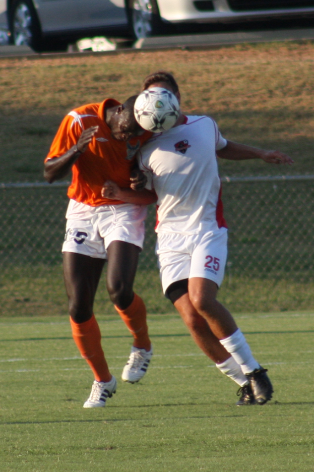 Zwei Spieler auf einem umzäunten Platz, einer im orangen T-Shirt, der den Ball mit dem Kopf trifft, mit Autos und Gras im Hintergrund.