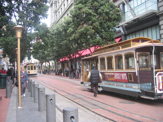 Ein Zug auf Schienen mit Text an der Seite, eine Person in der Nähe, kleine Pfosten mit Seilen links, Straßenlaternen, Bäume, viele Menschen, ein Müllcontainer links und Gebäude mit Leitern rechts.