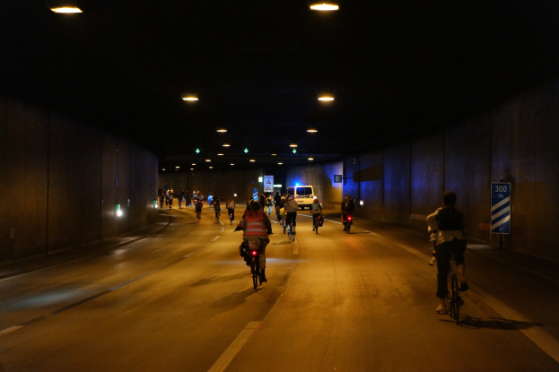 Eine Gruppe von Menschen, die nachts auf Fahrrädern eine Straße hinunterfahren, mit überkopfbeleuchtung, flankiert von Wänden und einem Fahrzeug im Hintergrund.