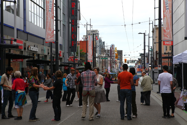 Eine Gruppe von Menschen, die sich an den H√§nden halten, steht auf einer Stra√e, die von Geb√§uden mit Gesch√§ften im Erdgeschoss, Stra√enlaternen und Masten mit Dr√∂hen und Plakaten ges√§umt ist; Zelte sind auf dem rechten Gehweg zu sehen.