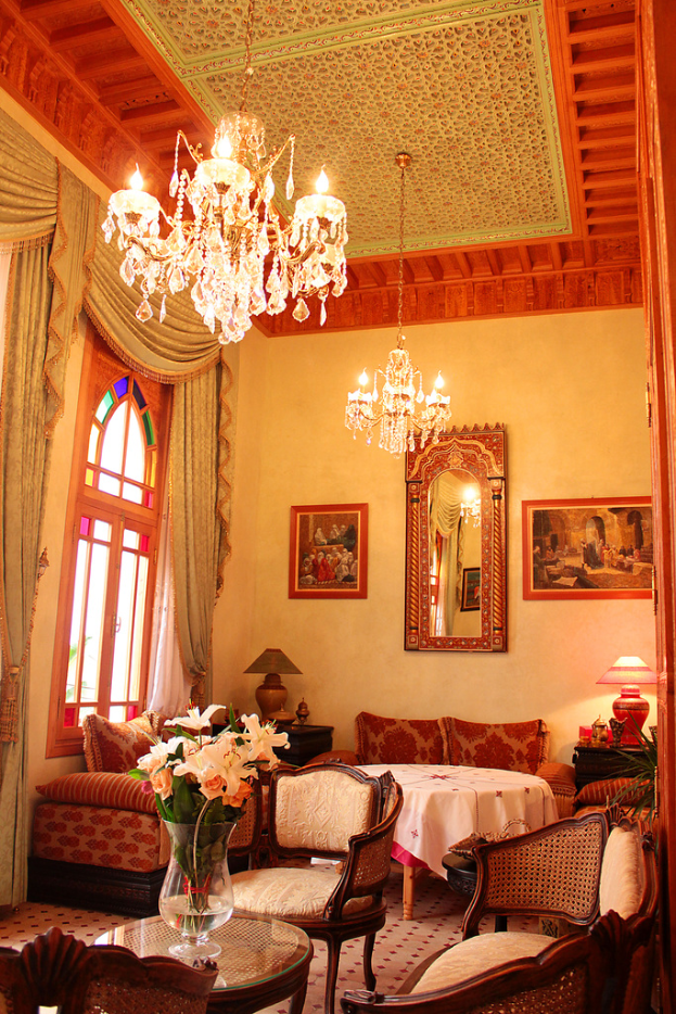 Ein Wohnzimmer mit einem Tisch und einem Blumentopf im Vordergrund, umgeben von Stühlen, Sofas und einem mit einer weißen Decke bedeckten Tisch im Hintergrund, gerahmte Bilder an der Wand, von der Decke hängende Lampen und Vorhänge mit einem Fenster links.