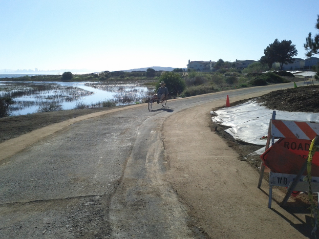 Ein Mann auf einem Fahrzeug auf einer Straße mit einer Barriere und einem Verkehrskegel auf der rechten Seite, mit Wasser, Bäumen, Gebäuden und Himmel im Hintergrund.