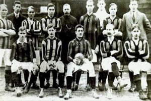 Schwarzes und weisses Foto einer Fussballmannschaft, einige stehen und einige sitzen, vor einer Wand mit dem Text "Ballymena Football Club" unten.