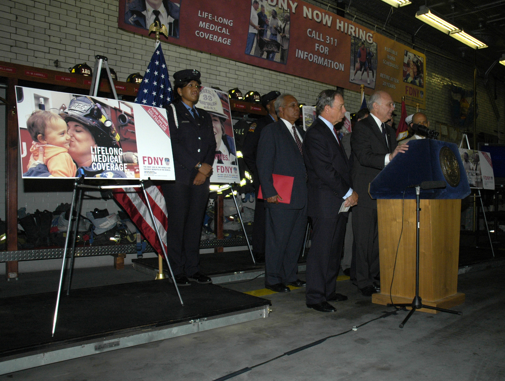 Eine Gruppe von Menschen steht in der Nähe eines Podiums mit Mikrofonen, Schildern auf Ständern, Fahnen und Schildern an der Wand, alles unter hellen Lichtern.
