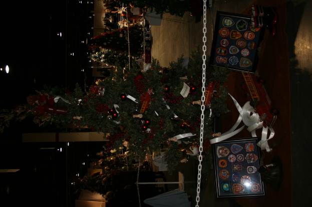 Ein Weihnachtsbaum mit Geschenken, Rahmen und Spielzeugen auf einer Plattform, mit Menschen, zus├Ątzlichen Weihnachtsb├Ąumen, Ketten, Pfosten und Lichtern im Hintergrund.