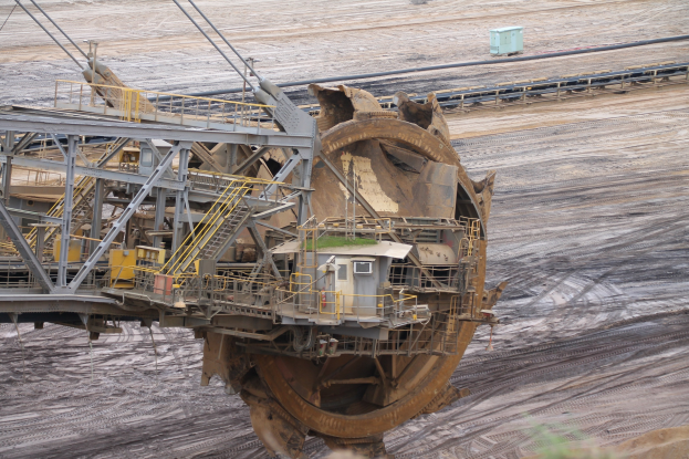 Großer Bagger auf einem Schmutzfeld mit Eisenstangen, Treppen, Geländern und anderen Gegenständen umgeben, umgeben von Blättern am Boden und Sand im Hintergrund, wahrscheinlich in einem Kohlebergwerk verwendet.