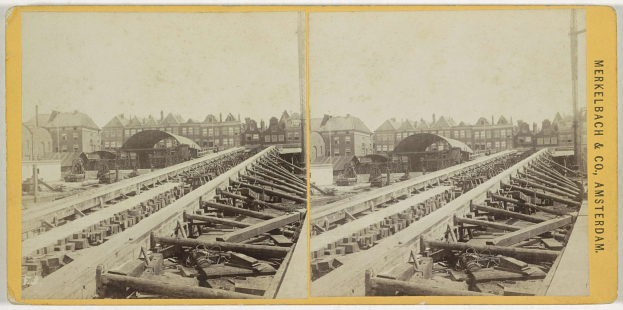 Schwarz-weiß-Foto einer Zugschiene mit Gebäuden im Hintergrund, Text 'Merkelbach & Co, Co. Amsterdam' rechts und hölzerne Gegenstände im Vordergrund verstreut.