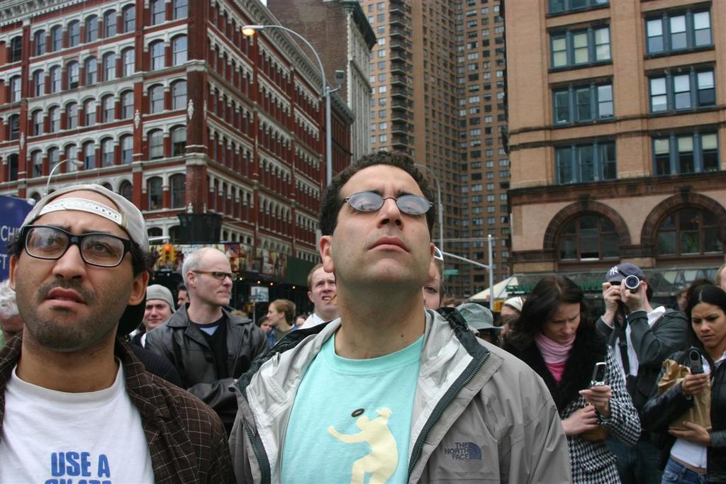 Mehrere Personen sind versammelt, wobei zwei Frauen Telefone halten und eine Person ein Objekt hält; Gebäude und Laternen sind im Hintergrund sichtbar.