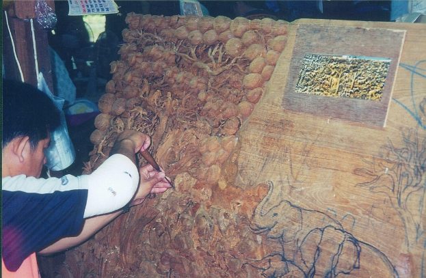Ein Mann schnitzt auf einer Holztafel mit einem Werkzeug, mit Darstellungen von Elefanten und Bäumen, einem Kalender, einer Tasche und einem Holzpfahl im Hintergrund.