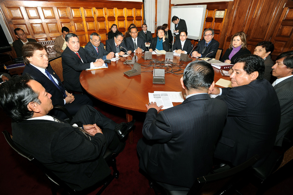 Eine Runde-Tisch-Sitzung mit Delegierten, die diskutieren, Mikrofone, Papiere, Kabel und Dateien auf dem Tisch, mit einer hölzernen Wand im Hintergrund.