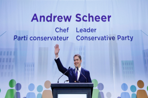 Andrew Scheer, Chef der konservativen Partei, steht hinter einem Podium mit zwei Mikrofonen und einem Bildschirm, mit einer Fahne im Hintergrund, die Text und Gebäudeabbildungen zeigt.