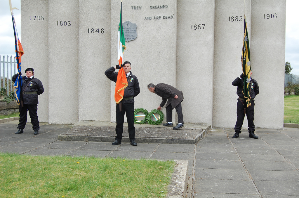 Drei uniformierte Personen halten Fahnen in verschiedenen Farben, während ein Mann hinter ihnen grüne Kränze hält, vor einer Wand oder Säulen mit Text, Gras, Gehweg, einem Zaun auf der linken Seite und Bäumen und einem Gebäude auf der rechten Seite.