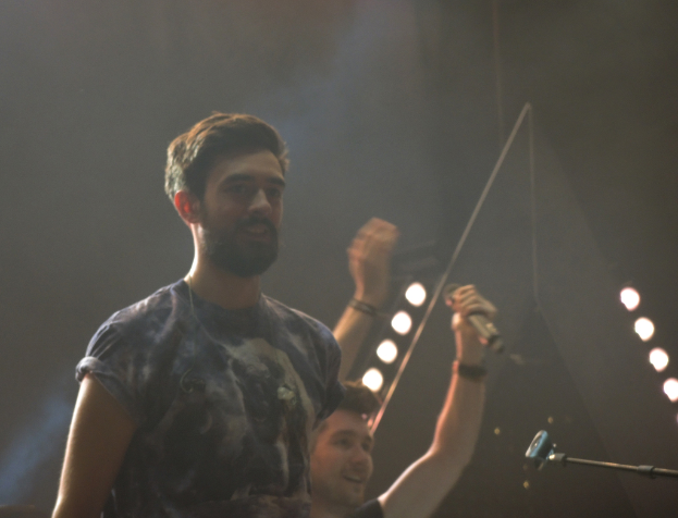 Ein Mann in einem T-Shirt lacht stehend mit einem anderen lachenden Mann, Lichtern, Rauch, einem Mikrofon und anderem Equipment im Hintergrund.