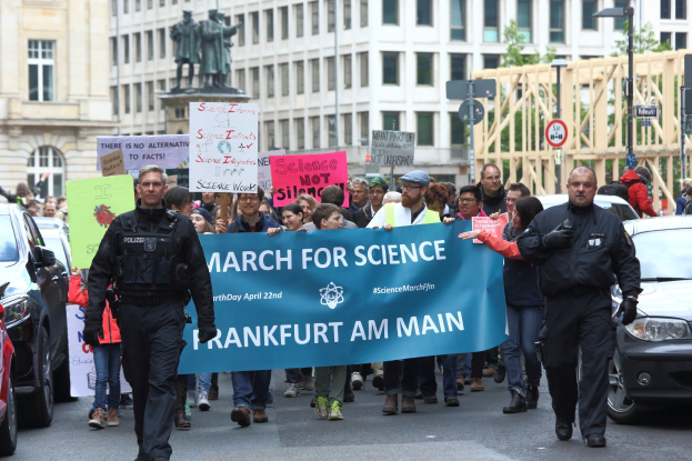 Gruppe von Menschen marschiert mit einem "March for Science Frankfurt am Main"-Schild die Straße entlang, Autos fahren daneben und Gebäude mit Fenstern, Statuen, Laternen, Schilder und Bäumen im Hintergrund.