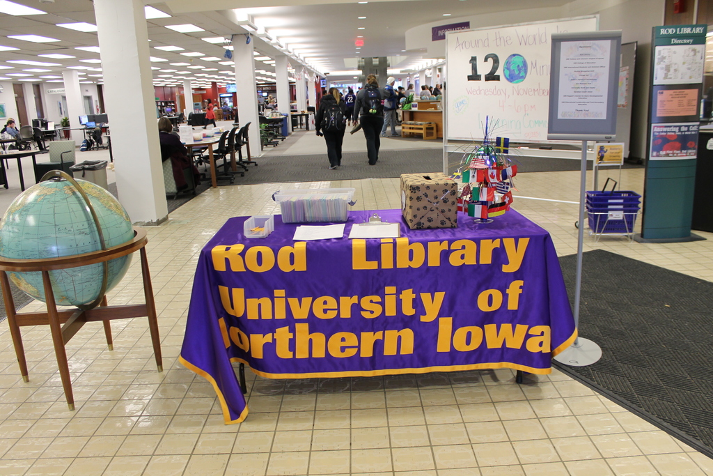 Eine Ausstellung mit einem Tisch, auf dem verschiedene Landesflaggen, eine Box, Papiere und eine Fahne sowie links daneben eine Weltkugel, rechts daneben eine Anschlagtafel mit einigen Menschen in der Nähe und im Hintergrund mehrere Tische und Stühle zu sehen sind.