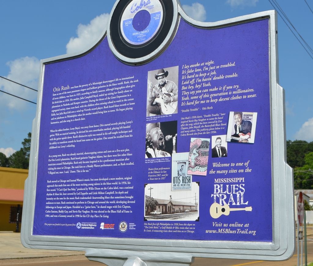 Ein Informationsbrett, Gebäude, ein Schuppen, Bäume, Stromkabel und ein bewölkter Himmel.