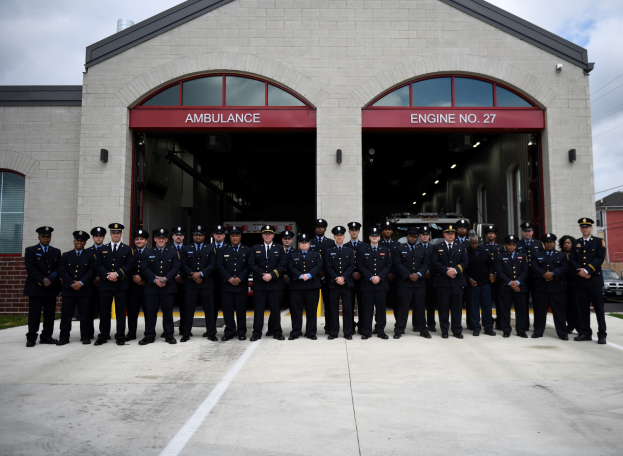 Feuerwehrleute in Uniform und Mützen vor einer Feuerwache mit Fahrzeugen und Gebäuden im Hintergrund unter einem klaren blauen Himmel.
