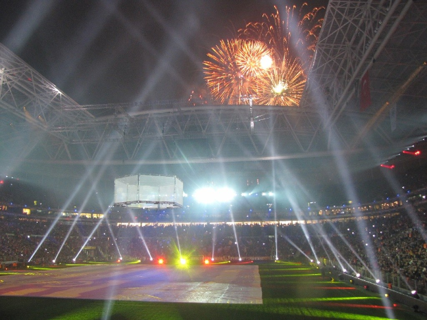 Das Olympische Stadion in London, England, bei Nacht beleuchtet mit einer Menge Menschen drumherum, grünem Rasen darunter und Feuerwerk am Himmel.