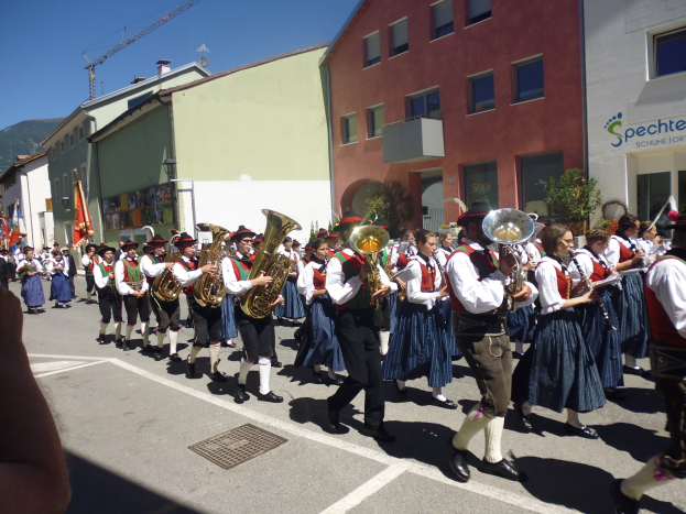 Gruppe von Menschen in traditioneller bayrischer Tracht, die Musikinstrumente spielen, während sie eine Straße mit Gebäuden, einigen Fahnen, einem Hügel und einem klaren blauen Himmel entlanggehen.