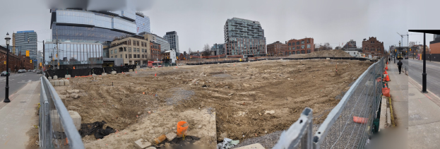 Eine Baustelle mit einem großen Haufen Erde in der Mitte, umgeben von Gebäuden, Straßeninfrastruktur, Fahrzeugen, Fußgängern und Bäumen unter einem klaren blauen Himmel.