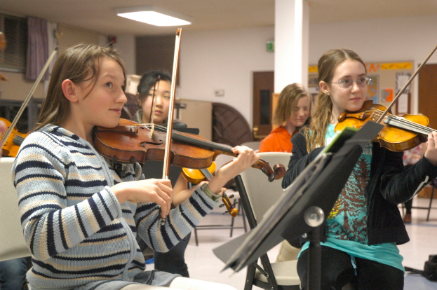 Eine Gruppe von Frauen, die auf Stühlen Geigen spielen, mit einem Buch auf einem Ständer, einer Tür, einer Säule, einem Vorhang und einem Fenster im Hintergrund.