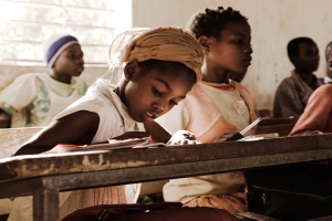 Eine Gruppe von Kindern sitzt an Tischen in einem Klassenzimmer mit Büchern und Taschen auf den Bänken, eine Wand und ein Fenster im Hintergrund.