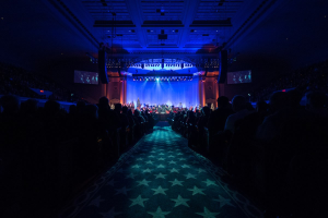 Ein großer Zuschauerraum mit Menschen auf Stühlen, eine Bühne mit Musikern und eine Decke mit Lichtern und Fernsehern, was auf eine Konzerthalle hinweist.