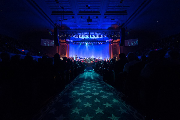 Ein großer Zuschauerraum mit Menschen auf Stühlen, eine Bühne mit Musikern und eine Decke mit Lichtern und Fernsehern, was auf eine Konzerthalle hinweist.