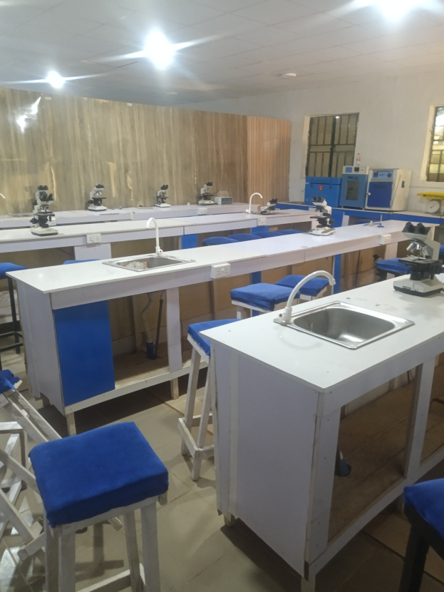 Ein Labor mit blauen Hockern, weißen Arbeitsplatten, einem Waschbecken, verschiedenen Maschinen, einer verglasten Wand und Deckenleuchten an der Universität für Wissenschaft und Technologie in Ahmedabad.