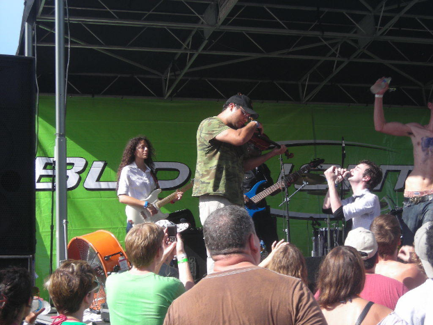 Eine Gruppe von Müssern tritt auf der Bühne mit verschiedenen Instrumenten auf, während das Publikum vor einem Podium steht und die Musik genießt. Ein großer schwarzer Lautsprecher ist links zu sehen, und ein grünes Banner ist im Hintergrund sichtbar.