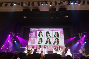 Eine Gruppe von Menschen, die auf einer hell erleuchteten Bühne performen, mit einer Menge darunter, mit einem Sänger mit Mikrofon und einem Musiker, der ein Instrument spielt, während ein großer Bildschirm Bilder der K-Pop-Gruppe BK48 anzeigt.