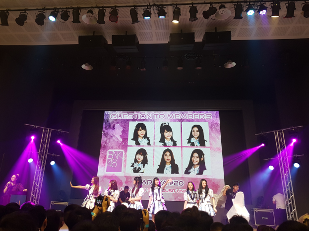 Eine Gruppe von Menschen, die auf einer hell erleuchteten Bühne performen, mit einer Menge darunter, mit einem Sänger mit Mikrofon und einem Musiker, der ein Instrument spielt, während ein großer Bildschirm Bilder der K-Pop-Gruppe BK48 anzeigt.