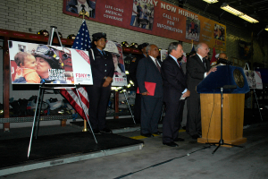 Eine Gruppe von Menschen steht neben einem Podium mit Mikrofonen, umgeben von Tafeln auf Ständern, Flaggen und Tafeln an der Wand, alles unter hellen Scheinwerfern.