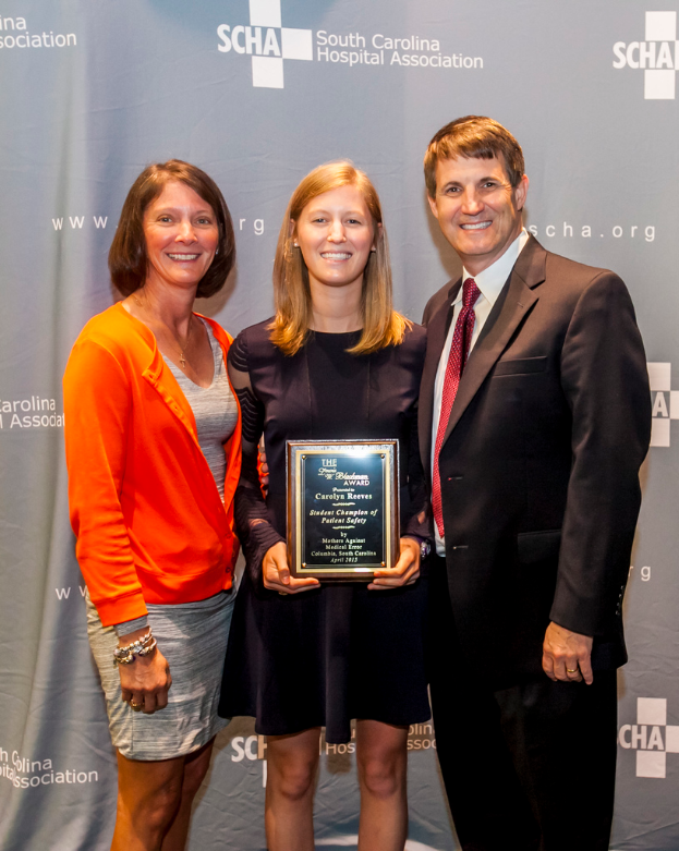 Drei Personen, zwei Frauen und ein Mann, stehen zusammen. Der Mann trägt einen schwarzen Anzug, eine Frau trägt ein orange-schwarzes Kleid und die andere Frau trägt ein schwarzes Kleid und hält eine Trophäe in der Hand.