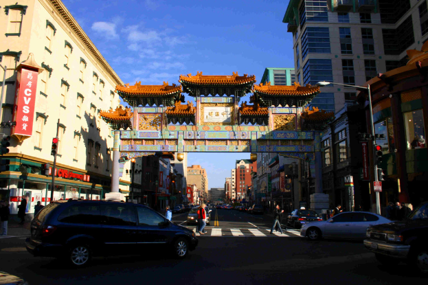 Eine Straße mit fahrenden Fahrzeugen, Fußgängern an einer Zebrastreifen, einem Bogen im Hintergrund, Gebäuden auf beiden Seiten und einer Ampel unter einem klaren Himmel.