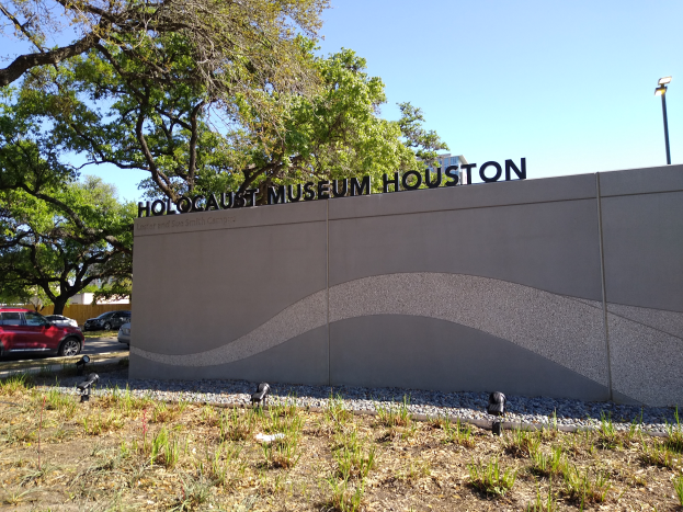 Das Holocaust-Museum in Houston, Texas, mit einer textbedeckten Wand, Steinen, Gras, Pflanzen, Bäumen, Fahrzeugen auf der Straße, einem Straßenpfahl und einem bewölkten Himmel.