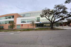 Großes, zweistöckiges Backsteingebäude mit der Aufschrift "Presbyte Mittelschule" und zahlreichen rechteckigen Fenstern, umgeben von Bäumen, Gras, Schildern, Strommasten und einer asphaltierten Straße unter einem bewölkten Himmel.