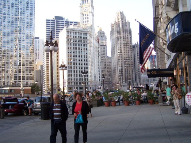 Eine belebte Straßenszene mit Menschen, Autos und Blumenkübeln, mit Gebäuden, Straßenlaternen, Geschäften und Fahnen im Hintergrund.