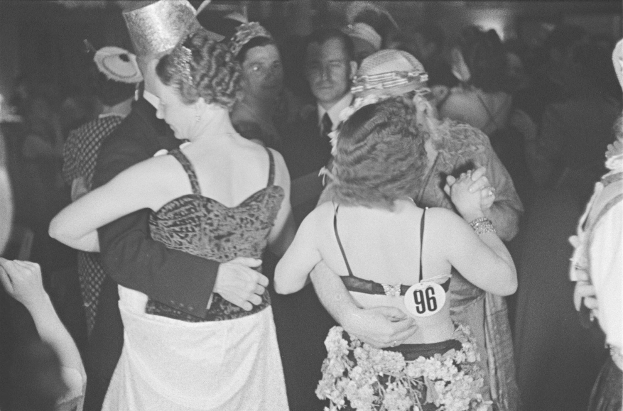 Schwarzes und weißes Foto einer Frau in einem Kleid und Hut, die auf einer Party tanzt und von anderen Menschen umgeben ist.