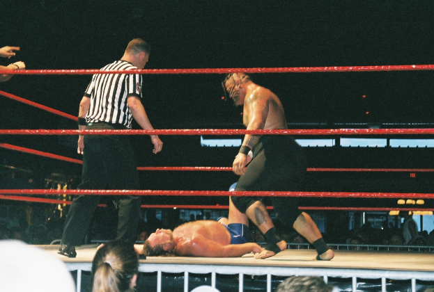 Zwei Boxer im Ring mit einem Mann in einem weiß-schwarzen gestreiftem T-Shirt daneben, mit roten Seilen im Vordergrund.