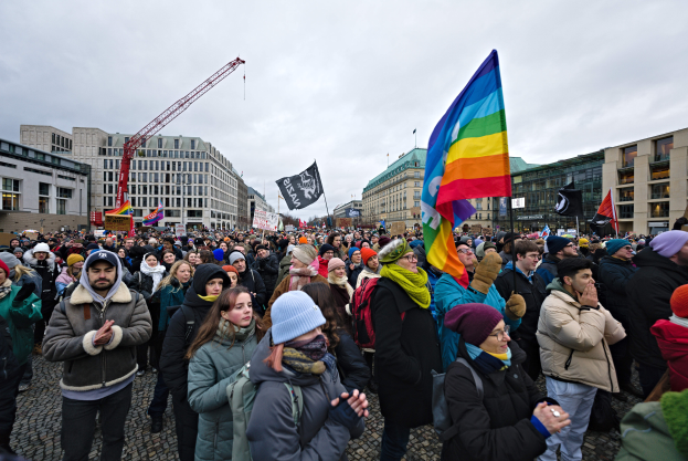 Eine große Gruppe von Menschen auf einer LGBTQ+-Rechtsmarsch in Berlin, die Fahnen und Plakate schwenken, mit Gebäuden, einem Kran und einem bewölkten Himmel im Hintergrund.