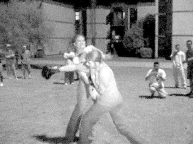 Zwei Personen boxen in der Mitte eines Schwarz-Weiß-Bildes, im Hintergrund ist ein Gebäude und einige Zuschauer zu sehen.