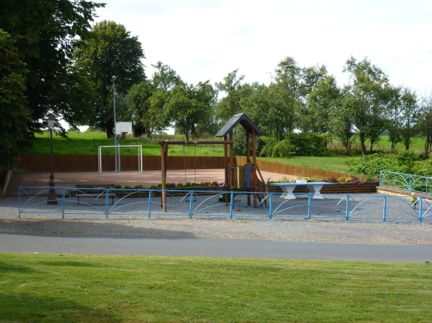 Park mit einem Spielplatz in der Mitte, umgeben von einer Straße, Gras, Pflanzen, Blumen, einem Zaun, Pfosten und einem Basketball-Korb, mit Bäumen und einem klaren blauen Himmel im Hintergrund.