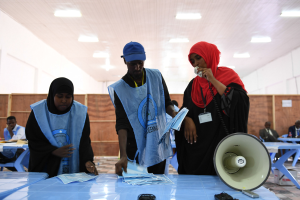 Eine Gruppe von Menschen steht um einen Tisch mit einem Megafon und Papieren an einer Wahllokal, während andere im Hintergrund sitzen und stehen, unter beleuchteter Decke.