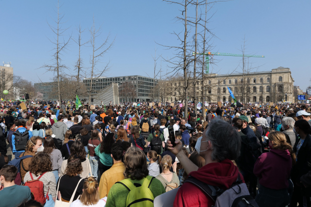 Große Menschenmenge mit Schildern und Taschen vor einem Gebäude mit Bäumen und wolkenlosem Himmel, was auf eine Klimaprotestveranstaltung hinweist.