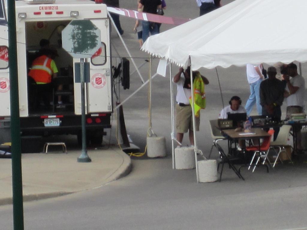Ein Rettungswagen steht links von einer Straße, während auf der rechten Seite ein Zelt viele Menschen schützt, mit einem Tisch und einem Stuhl in der Nähe.